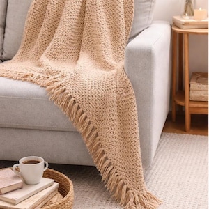 May include: A beige, textured throw blanket with fringe detail draped over a gray sofa. A woven basket with books and a coffee cup sits on the floor. The blanket's neutral color complements the room's decor.