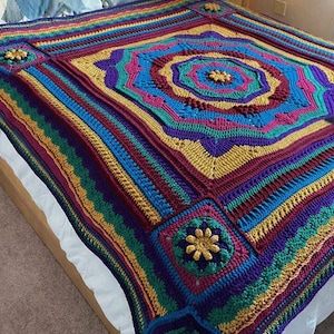 May include: A colorful, handmade crochet blanket with a geometric design. The blanket features a central medallion and corner squares, all adorned with flower-like embellishments. The color palette includes shades of blue, purple, yellow, and green.