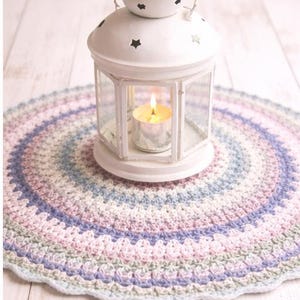 May include: A white metal lantern with a lit candle inside sits on a pastel-colored, crocheted doily. The lantern has a handle and star-shaped cutouts. The doily features concentric circles of blue, pink, and lavender.