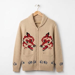 May include: A beige zip-up cardigan with two red and black wolf-like faces on the front. The sweater features a shawl collar, two front pockets, and paw print designs near the bottom. It is displayed on a wooden hanger.