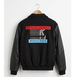 May include: Black bomber jacket with black leather sleeves. The back of the jacket features a graphic with the text "MICHAEL JACKSON" in red, a stylized image of a dancer, and the text "WORLD TOUR" in blue.