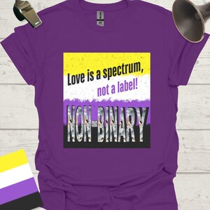 May include: Purple t-shirt with the text "Love is a spectrum, not a label!" above the word "NON-BINARY" in a stylized font. The design incorporates the colors of the non-binary pride flag: yellow, white, purple, and black.
