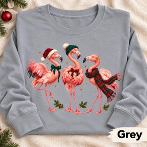 May include: A grey crewneck sweatshirt featuring a graphic of three pink flamingos. One flamingo wears a red Santa hat and green bow, the second wears a green knit hat and holds a gift, and the third wears a red and green plaid scarf.