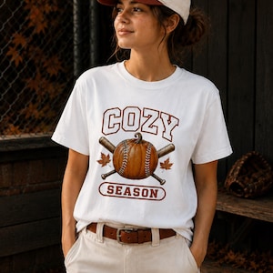 May include: A white t-shirt featuring a graphic of a pumpkin designed like a baseball, crossed baseball bats, and autumn leaves, with the text COZY SEASON. The shirt is paired with a white baseball cap with a brown brim and a brown leather belt.