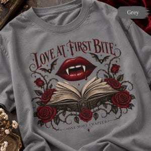 May include: A grey t-shirt featuring a graphic print of red vampire lips with fangs, an open book, and red roses. The text "LOVE AT FIRST BITE" is arched above the lips, and "ONE MORE CHAPTER" is printed below the book, surrounded by small bats.