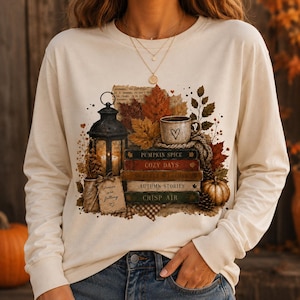 Pode incluir: Uma camisola de manga comprida cor de creme com uma impressão gráfica de uma lanterna preta, uma caneca com um coração e uma pilha de livros intitulados Pumpkin Spice, Cozy Days, Sweater Weather, Autumn Stories e Crisp Air.