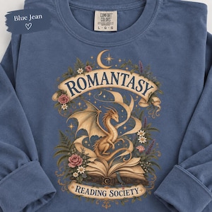 May include: A blue jean colored long-sleeve shirt featuring a graphic of a dragon emerging from an open book. The text reads ROMANTASY and READING SOCIETY. The design includes floral elements, a crescent moon, and stars in muted earth tones.