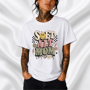May include: A white short-sleeved t-shirt featuring a graphic design with the text "SOFT ALT MOM" in a distressed font. The design includes a yellow melting smiley face, pink and green flowers, black and white checkerboard patterns, and stars.
