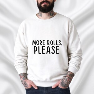 Op de afbeelding: Wit sweatshirt met de tekst "MORE ROLLS, PLEASE" in zwarte blokletters. Het sweatshirt heeft een ronde hals en lijkt van een zacht materiaal te zijn. Het thema is vrijetijdskleding.