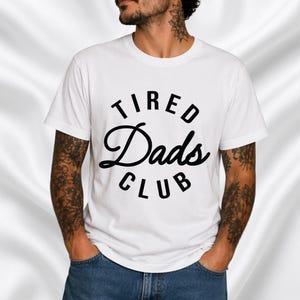 Op de afbeelding: Wit T-shirt met de tekst "TIRED Dads CLUB" in zwarte, vette en gestileerde letters. De tekst is in een cirkel geplaatst. Het T-shirt heeft een ronde hals.