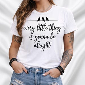 Op de afbeelding: Wit T-shirt met de tekst "every little thing is gonna be alright" in zwarte letters. Boven de tekst staan drie zwarte vogel silhouetten. Een casual top voor dagelijks gebruik met een positieve boodschap.