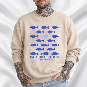 Op de afbeelding: Beige sweatshirt met een blauwe visafbeelding. Het ontwerp bevat rijen blauwe vissen, met één vis in een lichtere blauwtint. De tekst "CALLED TO BE DIFFERENT ROMANS 12:2" is eronder gedrukt.