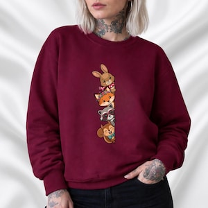 Op de afbeelding: Bordeaux sweatshirt met een verticaal ontwerp van cartoon dieren: een konijn, een vos, een wasbeer en een eekhoorn. Het konijn draagt een sjaal, de vos een bloemenkroon, de wasbeer een zonnebril en de eekhoorn een vlinderdas.
