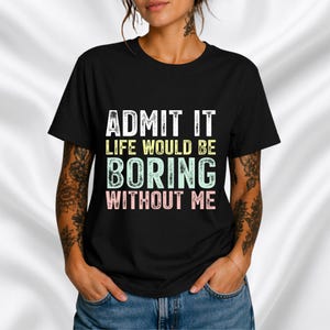 Op de afbeelding: Zwart T-shirt met de tekst "GEEF HET TOE, HET LEVEN ZOU ZONDER MIJ VERVELEND ZIJN" in wit, geel, groen en roze. Het T-shirt heeft een ronde hals en wordt gedragen.