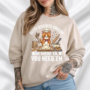 Op de afbeelding: Beige sweatshirt met een cartoon kattenkok in een bakkerij. De kat draagt een koksmuts en een rode vlinderdas. Tekst luidt "Kitty Biscuits Bakery" en "We Knead 'Em, You Need 'Em."