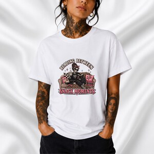 Może przedstawiać: Biały t-shirt z grafiką przedstawiającą szkielet siedzący na kanapie w panterkę, czytający książkę z kubkiem, przekąskami i poduszkami. Napis głosi READING BETWEEN SNACK MOMENTS czarnymi i różowymi literami.