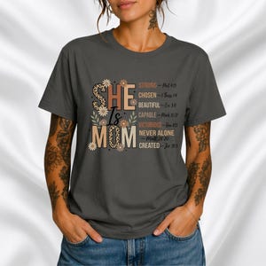 Op de afbeelding: Donkergrijs t-shirt met de tekst "SHE Is MOM" in een bloemen- en luipaardprint. Het shirt heeft ook een lijst met inspirerende woorden en bijbelverzen aan de rechterkant.