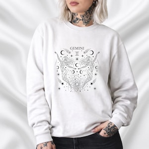 Op de afbeelding: Witte sweatshirt met een zwart Gemini dierenriem ontwerp. De afbeelding bevat twee gestileerde figuren, halve manen, sterren en het woord "Gemini". Het ontwerp is gecentreerd op de voorkant van de sweatshirt.