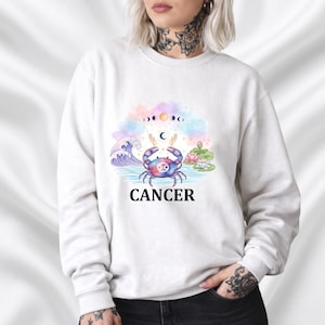 Op de afbeelding: Witte sweatshirt met een Kreeft sterrenbeeld ontwerp. De afbeelding toont een krab, golven, maanfasen en het woord "CANCER" in het zwart. Het ontwerp is in aquarel stijl met pastelkleuren.