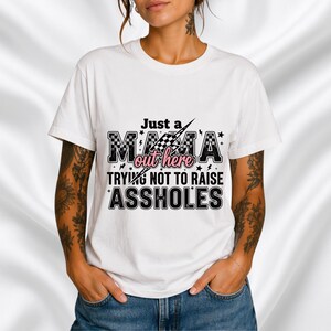 Op de afbeelding: Wit T-shirt met de tekst "Just a MAMA out here TRYING NOT TO RAISE ASSHOLES" in zwart en roze. Het ontwerp bevat een geblokte vlag en sterren.
