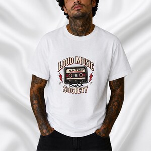 May include: A white short-sleeved t-shirt featuring a graphic of a black cassette tape with the text "PLAY IT LOUD" on the tape, surrounded by the words "LOUD MUSIC SOCIETY" in a curved font, accented by red lightning bolts and small stars.