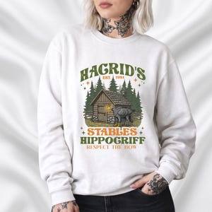 Op de afbeelding: Witte sweatshirt met een grafisch ontwerp. Het ontwerp toont "Hagrid's Stables" met een hut, bomen en een hippogrief. De tekst bevat "EST. 1981" en "Respect the Bow".