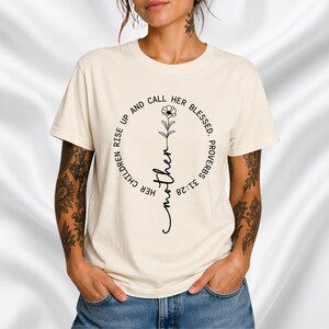 Op de afbeelding: Crèmekleurig T-shirt met een cirkelvormig ontwerp. Het ontwerp bevat het woord "Mother" in cursief, een bloemenillustratie en de tekst "Her children rise up and call her blessed. Proverbs 31:28."