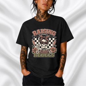 Op de afbeelding: Een zwart t-shirt met een grafische print van een skelet met een zonnebril en een hoofdband met luipaardprint. Het ontwerp bevat een zwart-witte geblokte achtergrond, roze bloemen, bliksemschichten en de tekst RAISING little REBELS.