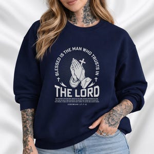 Op de afbeelding: Marineblauwe sweatshirt met de tekst "Gezegend is de man die op de Heer vertrouwt." Het ontwerp bevat biddende handen, een kruis en de Bijbeltekst Jeremia 17:7-8. De sweatshirt is effen van kleur.
