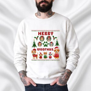 Op de afbeelding: Witte sweatshirt met een feestelijk "Merry Woofmas" ontwerp. Het ontwerp toont verschillende honden met kerstmutsen en truien, evenals kerstbomen en pootafdrukken. De tekst is in rood en groen.