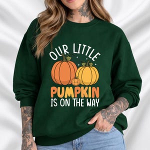 Op de afbeelding: Donkergroene sweatshirt met de tekst "OUR LITTLE PUMPKIN IS ON THE WAY" in wit en oranje. Het ontwerp toont drie pompoenen in oranje en geel. Een herfstthema-afbeelding voor aanstaande ouders.