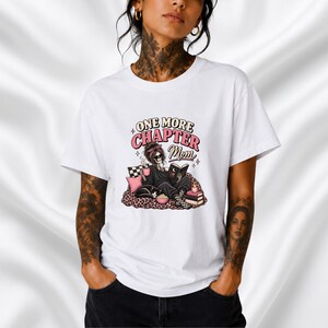 Op de afbeelding: Een wit t-shirt met korte mouwen en een grafische print van een skelet dat een boek leest met de tekst "ONE MORE CHAPTER Mom". Het skelet draagt een hoofdband en een zonnebril, omringd door een kat, een kom popcorn en boeken.