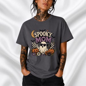 Op de afbeelding: Een donkergrijs t-shirt met een grafische print met de tekst "SPOOKY MOM". Het ontwerp bevat een geest met een hartvormige zonnebril, twee oranje pompoenen, een maansikkel, vleermuizen, bloemen en een klein hartvormig bordje met de tekst "RAISING LITTLE MONSTERS".