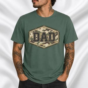 Op de afbeelding: Groen t-shirt met een camouflage ontwerp in een ruitvorm, met het woord "DAD" in een vet, vintage lettertype. Het shirt is effen groen van kleur en heeft korte mouwen.