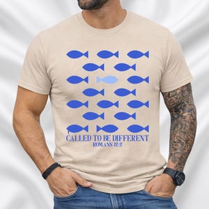 Op de afbeelding: Beige T-shirt met een blauw vispatroon en één lichtere blauwe vis. De tekst "CALLED TO BE DIFFERENT ROMANS 12:2" is onder het visontwerp gedrukt. Het shirt is gemaakt van een zacht materiaal.