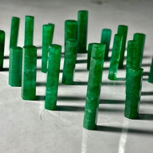 Op de afbeelding: Een verzameling heldergroene smaragdkristallen, elk met een rechthoekige vorm en facetranden. De kristallen zijn gerangschikt op een lichtgekleurd oppervlak, waardoor een display van natuurlijke edelstenen ontstaat. De smaragden variëren in hoogte en zijn verlicht om hun kleur te benadrukken.