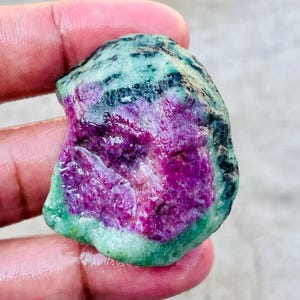 Puede incluir: Una gema en bruto con una vibrante exhibición de colores. La piedra presenta un centro magenta intenso, rodeado por una banda verde y una capa exterior de color verde azulado oscuro. La textura natural y las variaciones de color crean una apariencia única.