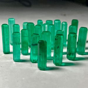 Op de afbeelding: Een verzameling smaragd edelstenen, elk met een gefacetteerde, cilindrische vorm. De edelstenen hebben een levendige groene kleur en zijn gerangschikt op een wit oppervlak. De edelstenen variëren enigszins in grootte en helderheid.