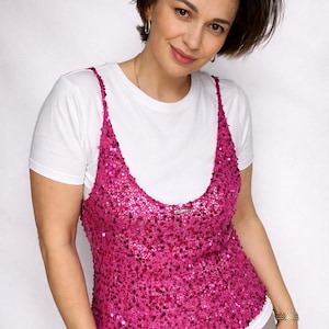 Op de afbeelding: Een vrouw draagt een wit t-shirt onder een felroze pailletten top. De top heeft dunne bandjes en een ronde halslijn. De vrouw draagt ook gouden sieraden en een beige broek. De achtergrond is effen wit.