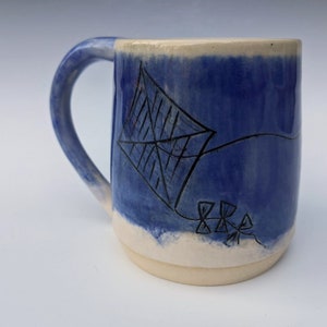 May include: A blue ceramic mug with a white rim and handle. The mug has a black kite design with a string and three small flowers.