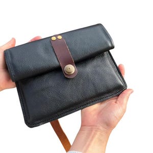 Puede incluir: Un bolso de mano de cuero negro con cierre de solapa, asegurado por una correa de cuero marrón y un botón de latón. El bolso se sostiene abierto, mostrando su interior. El cuero tiene una textura.