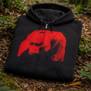 Puede incluir: Sudadera con capucha negra con cremallera y un gran gráfico rojo de la cara de un primate. La sudadera tiene una capucha con cordón y un bolsillo de canguro. La imagen está sobre un suelo forestal con musgo y hojas.