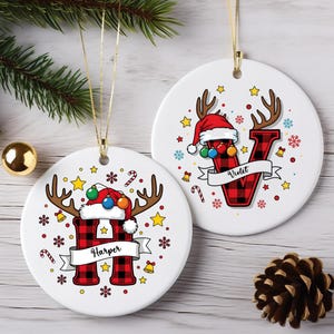 May include: Two round white ceramic ornaments featuring red and black buffalo plaid letters H and V. Each letter is topped with a Santa hat, reindeer antlers, and colorful ornaments. A white banner displays the names Harper and Violet.