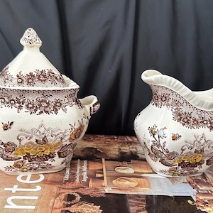 Può includere: Una zuccheriera in ceramica color crema con coperchio e una lattiera abbinata. Entrambi i pezzi presentano un dettagliato motivo floreale marrone a trasferimento con accenti di giallo e arancione, inclusi disegni di fiori e volute ornamentali.