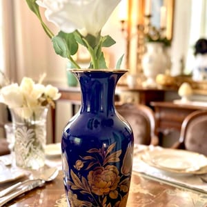 Könnte beinhalten: Eine tief kobaltblaue Keramikvase mit einem goldfarbenen Blumenmuster aus Rosen und Blättern am Körper. Die Vase hat einen ausgestellten Rand mit einem dünnen goldenen Besatz und enthält eine einzelne weiße Blume mit grünen Stielen und Blättern.