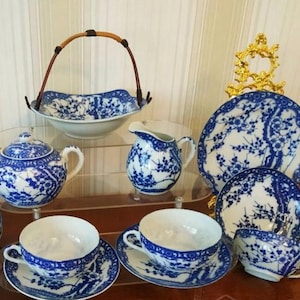 May include: A collection of white porcelain tableware featuring a blue floral pattern. The set includes a lidded sugar bowl, a creamer, two teacups with saucers, a small bowl, a large plate, and a bowl with a brown handle, all displayed on tiered stands.