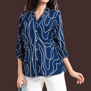 May include: A navy blue button-down shirt with a white, abstract, wavy pattern. The shirt has three-quarter sleeves and a collared neckline. The model is wearing white pants. The shirt is a stylish addition to any wardrobe.