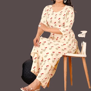 Könnte beinhalten: Cremefarbenes Outfit mit Blumenmuster, bestehend aus einem langärmeligen Oberteil und einer passenden Hose. Das Outfit hat ein rotes Kirschblütenmuster. Das Model trägt transparente Sandalen und sitzt auf einem weißen Stuhl mit Holzbeinen.