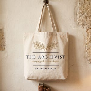 Könnte beinhalten: Eine cremefarbene Stofftasche aus Segeltuch mit zwei Schulterriemen. Auf der Vorderseite befindet sich eine goldene Lorbeerzweiggrafik über dem Text THE ARCHIVIST, dem Satz carrying what came before und dem Namen VALDREN HOUSE in dunkelblauer Schrift.