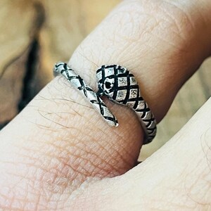 May include: A silver snake ring with a detailed, textured design. The snake's head features a diamond pattern, and the body wraps around the finger. The ring is a unique piece of jewellery.